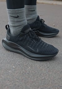 Zwarte sportschoenen met een gestructureerde bovenkant, voorzien van een subtiel patroon, zwarte veters en logo. Gecombineerd met geribde grijze sokken.