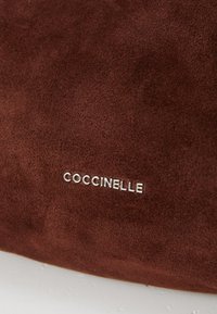 Smeđa suede torba s glatkom teksturom, koja sadrži srebrni logo "Coccinelle". Dizajn je minimalistički i ne uključuje dodatne ukrase.