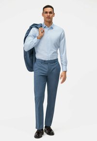 Lichtblauw overhemden met knoopsluiting, gecombineerd met getailleerde donkerblauwe broek met structuur en zwarte schoenen, jasje over de schouder.