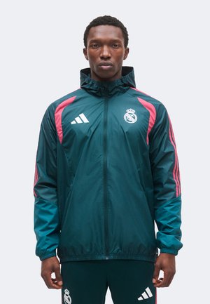 Muž oblečený v tyrkysovo-ružovej teplákovej bunde a nohaviciach Real Madrid Adidas, stojaci pred jednoduchým svetlým pozadím.