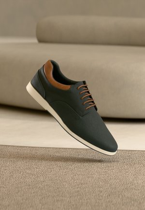 Chaussure décontractée noire à lacets avec des accents marron et une semelle blanche flottant au-dessus d'un tapis beige texturé devant un canapé beige.