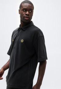 Νεαρός άνδρας που φοράει μαύρο κοντομάνικο πόλο με κίτρινα λογότυπα FC Bayern Munich και Adidas, κοιτάζει αριστερά του μπροστά σε απλό φόντο.