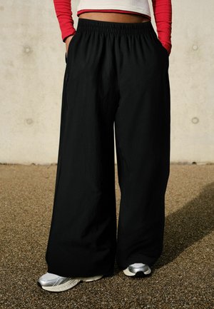 Person iført løstsiddende sorte bukser med vide ben, sølvfarvede sneakers og en hvid cropped top med røde lange ærmer, stående på grusjord.
