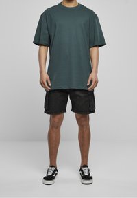 Persona che indossa una maglietta oversize verde scuro, pantaloncini cargo neri e sneakers basse nere con lacci bianchi, in piedi contro uno sfondo neutro.