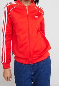 Červená bunda na zip z hladké látky, s bílými pruhy na ramenou a logem Adidas na hrudi. Kapsy po stranách.