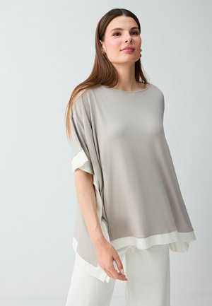 Giovane donna con lunghi capelli castani indossa una maglia larga grigio chiaro con bordo bianco e pantaloni bianchi, in piedi contro uno sfondo chiaro uniforme.