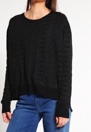 Femme portant un pull noir texturé à manches longues et un jean bleu, debout devant un fond blanc uni.