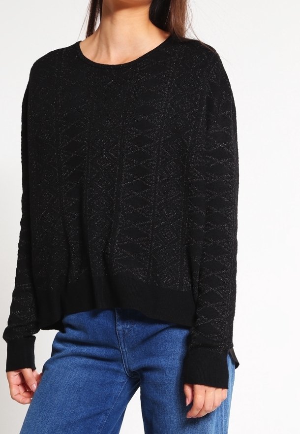 Femme portant un pull noir texturé à manches longues et un jean bleu, debout devant un fond blanc uni.