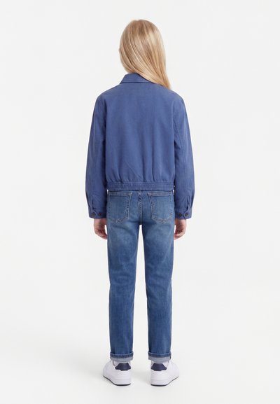 Νεαρό άτομο με μακριά ξανθά μαλλιά, που φοράει μπλε μπουφάν, τζιν παντελόνι με γυρισμένα μπατζάκια και λευκά αθλητικά παπούτσια, στέκεται με την πλάτη προς τα εμπρός σε λευκό φόντο.