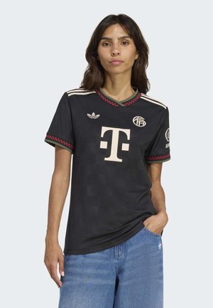 Woman wearing black soccer jersey with white Telekom logo, Adidas and team badges, paired with blue jeans, standing with one hand in pocket.