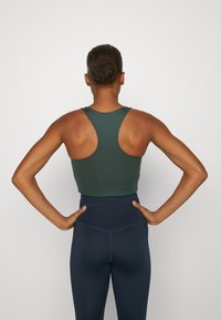 Zeleni bezrukavni crop top s dizajnom racerback, uparen s hlačama visokog struka u tamnoplavoj boji, koje prikazuju glatku teksturu i prilagođeni stil.