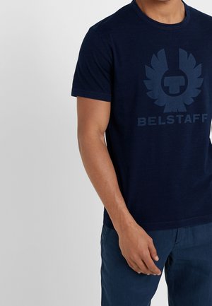 Homme portant un t-shirt Belstaff bleu foncé et un pantalon bleu assorti, debout devant un fond gris clair uni.
