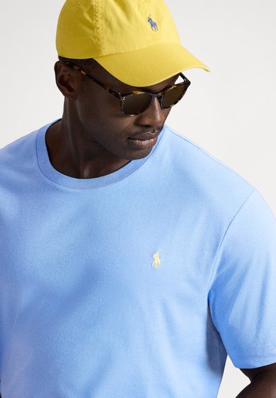 Šviesiai mėlynas marškinėliai su apvalia iškirpte ir geltonu siuvinėtu polo logotipu. Derinamas su geltona kepure ir tamsiais saulės akiniais.