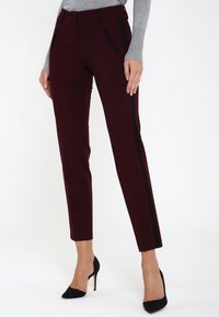 Pantalons taillés bordeaux avec des rayures noires sur les côtés, présentant une texture lisse et une coupe slim. La taille comprend des passants de ceinture et des poches.