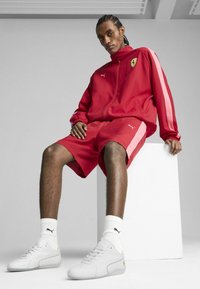 Chándal rojo de Puma con acentos en rosa en bloques de color, combinado con zapatillas blancas. Modelo sentado sobre un bloque blanco, mostrando unos pantalones cortos de corte relajado.