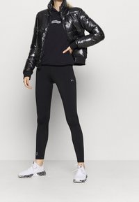 Svart puffjacka med blank yta, svart hoodie med logotyp, svarta leggings och vita sneakers med mönstrade sulor.