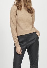 Light brown sweater with long sleeves and a round neckline, made of soft, textured material, paired with black, shiny tapered pants.
