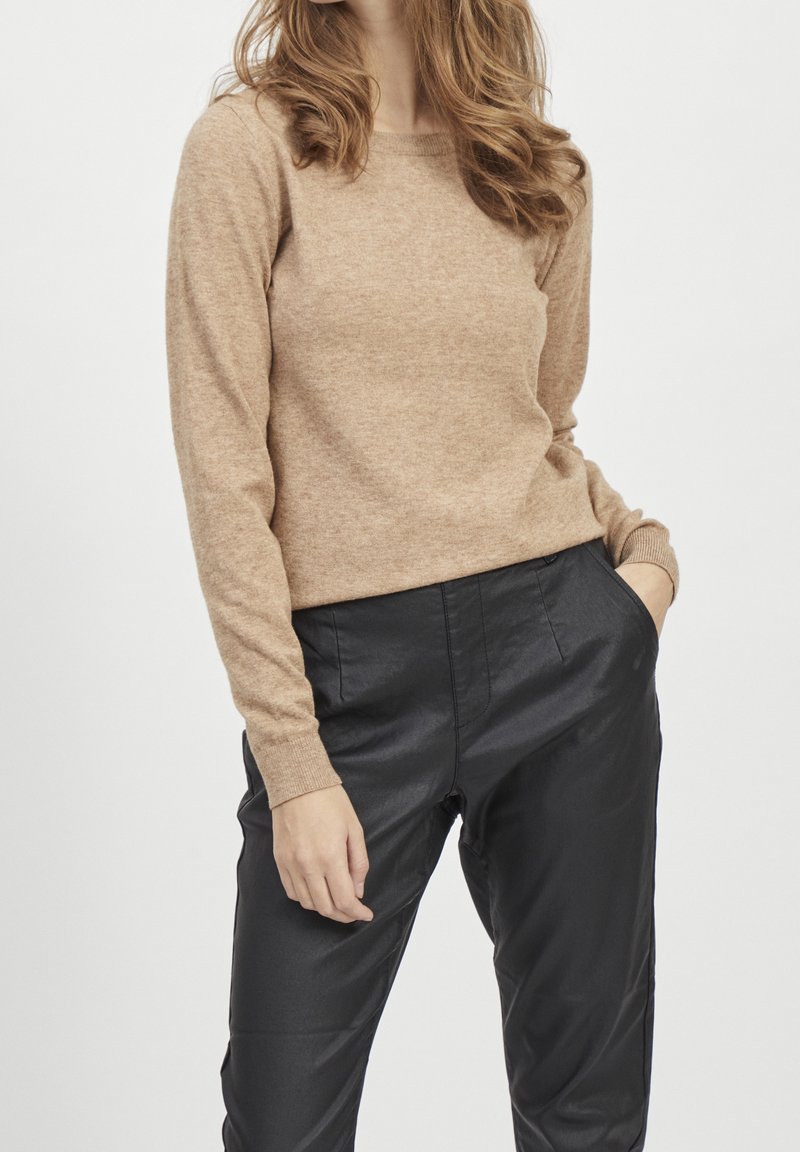 Light brown sweater with long sleeves and a round neckline, made of soft, textured material, paired with black, shiny tapered pants.