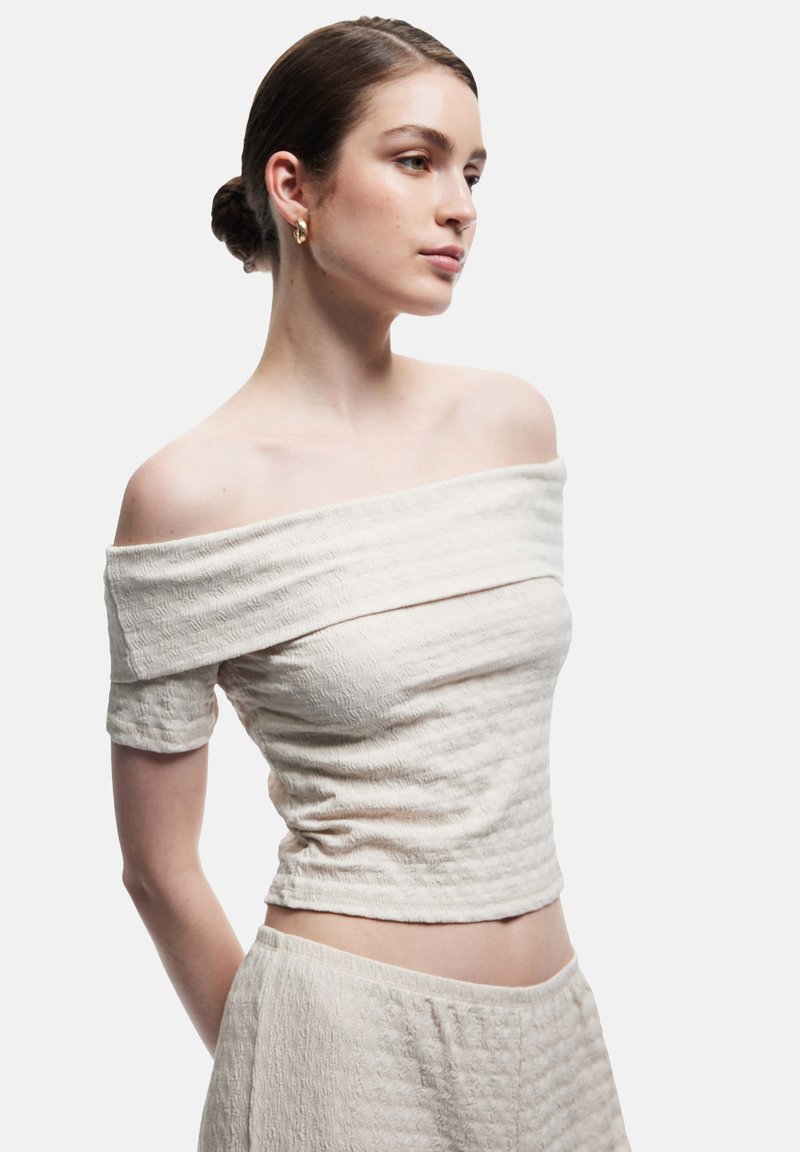 Top de color beige claro con los hombros al descubierto, fabricado con un tejido texturizado y arrugado. Diseño ajustado, mangas cortas y largo recortado.