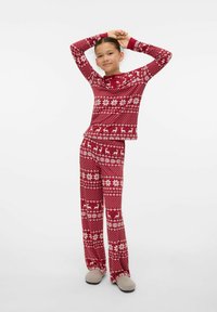 Młoda osoba stojąca z rękami na głowie, ubrana w czerwone piżamy świąteczne z białymi płatkami śniegu i reniferami, oraz szare kapcie.