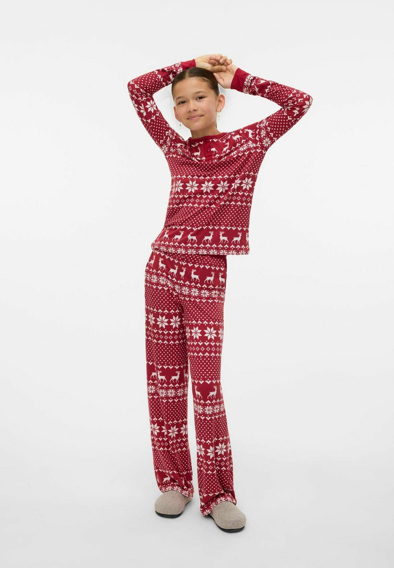 Młoda osoba stojąca z rękami na głowie, ubrana w czerwone piżamy świąteczne z białymi płatkami śniegu i reniferami, oraz szare kapcie.