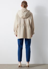 Beżowa kurtka z kapturem, z talią ściągniętą, długimi rękawami i gładką fakturą; zestawiona z niebieskimi dopasowanymi dżinsami i jasnymi obcasami.
