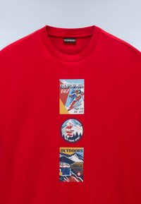 Rød bomulds t-shirt med tre grafiske patches: en skiløber, en skilift og en bjerglodge, alle med retro design elementer.