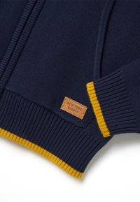 Marineblauer Strickcardigan mit gelben gerippten Bündchen, versehen mit einem Lederpatch mit der Aufschrift "NEW YORK VARSITY" an der Seite. Glatte Textur.