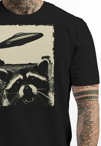 Schwarzes T-Shirt mit einem grafischen Druck eines Waschbären und einem UFO im Vintage-Stil, vor einem kontrastierenden cremefarbenen Hintergrund.