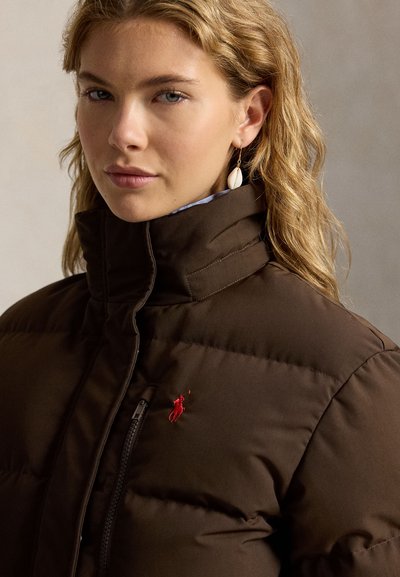 Femme aux cheveux blonds ondulés portant une doudoune marron avec un logo rouge brodé et une boucle d’oreille en coquillage à l’oreille gauche.