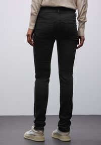 Jeans neri slim-fit con una texture liscia, dotati di tasche posteriori e vita a media altezza, abbinati a scarpe casual di colore chiaro.