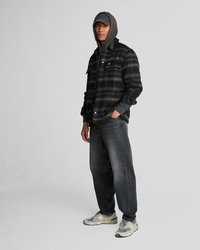 Veste à carreaux noirs avec des accents jaunes, sweat à capuche gris en dessous, jeans gris et baskets grises avec des accents verts. Le modèle se tient avec une main sur la hanche.