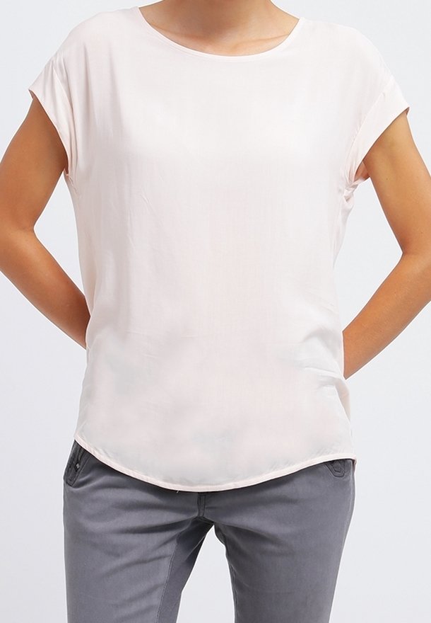 Haut rose clair à manches courtes en tissu lisse et léger, avec un col rond et un ourlet légèrement arrondi. Pantalon gris visible en dessous.