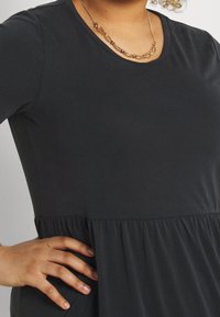 Robe noire à manches courtes en tissu doux avec un corsage ajusté et une taille évasée froncée. Un collier en chaîne dorée met en valeur le décolleté.