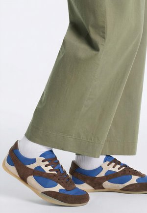 Een paar blauwe, bruine en beige sneakers gedragen met witte sokken en olijfgroene broek, zijaanzicht tegen een witte achtergrond.