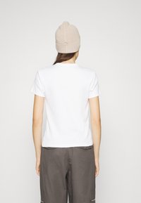 Bílé krátké tričko s kulatým výstřihem, nošené s šedými cargo kalhotami, zobrazené zezadu. Čepice je béžová pletená.