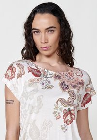 Blouse blanche en jersey avec des motifs floraux et paisley colorés. Présente un col bateau et des manches courtes. Texture lisse, coupe décontractée.