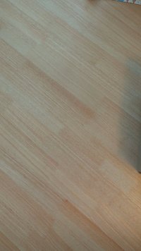 Superficie de madera clara con una textura suave, que presenta un patrón de grano natural y variaciones de color que van desde un beige pálido hasta un marrón claro.