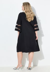 Eine Frau mit langen Haaren dreht sich leicht, trägt ein schwarzes Kleid mit durchsichtigen gestreiften Ärmeln und silbernen High-Heels, vor einem weißen Hintergrund.