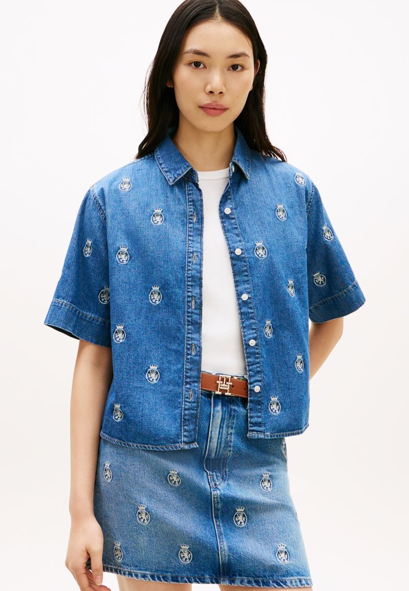 Νεαρή γυναίκα που φοράει ταιριαστή μπλε τζιν κοντομάνικη πουκαμίσα και φούστα με μικρά μοτίβα κορώνας και θυρεού, λευκή μπλούζα και καφέ ζώνη.