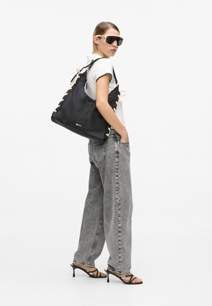 Γυναίκα που φοράει πολύ φαρδιά γκρι τζιν, λευκό πόλο, μαύρα σανδάλια με ψηλό τακούνι, μεγάλα γυαλιά ηλίου και κρατάει μια μεγάλη μαύρη τσάντα ώμου.