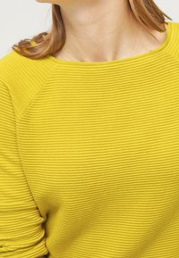 Haut en tissu côtelé jaune avec un col rond et des manches raglan, présentant une surface texturée et une finition douce.