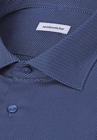 Gros plan d'une chemise à rayures bleues avec col, un bouton et une étiquette blanche portant la mention "seidensticker" à l'intérieur du col.