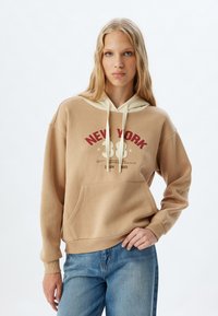 Sudadera beige con cordón color crema; presenta un gráfico de "NUEVA YORK", bolsillo tipo canguro y puños de canalé. Combinada con jeans de mezclilla azul.