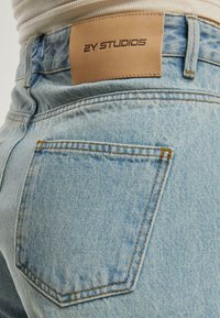 Jeans din denim albastru deschis cu o etichetă din piele pe care scrie "2Y STUDIOS." Prezintă un buzunar pe spate cu cusături contrastante și o textură decolorată.