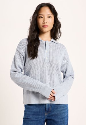 Frau mit langen dunklen Haaren trägt einen hellblauen Strickpullover mit Knöpfen und blaue Jeans, steht mit vor dem Körper verschränkten Händen.