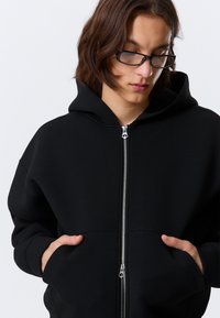 Hanorac negru cu glugă și fermoar în față, confecționat dintr-un material moale. Dispune de buzunare tip cangur și are un design cu croială lejeră.