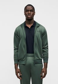 Πράσινο αθλητικό μπουφάν με φερμουάρ και ριμπ μανσέτες, συνδυασμένο με παντελόνι στο ίδιο χρώμα. Από κάτω, μια σκούρα πόλο μπλούζα. Ομαλή υφή με λεπτομέρειες λογοτύπου.