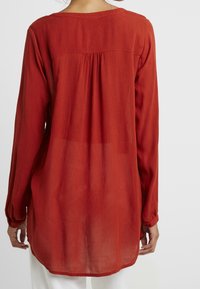 Blouse rouge à manches longues en tissu texturé, avec un col arrondi et un pli cousu au dos, tombant sous les hanches.