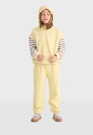 Enfant portant un sweat à capuche jaune pâle, un t-shirt à manches longues rayé en dessous, un pantalon de survêtement assorti et des baskets rayées, debout les yeux fermés.